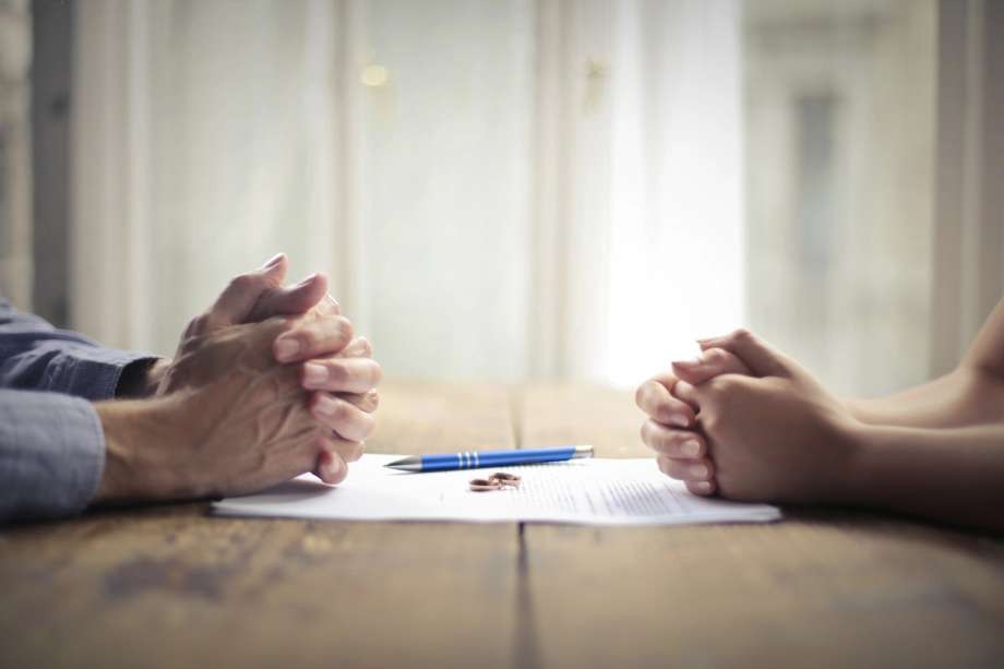 A close-up of two individuals sitting opposite each other at a wooden desk, their hands clasped while discussing legal paperwork or a settlement agreement.