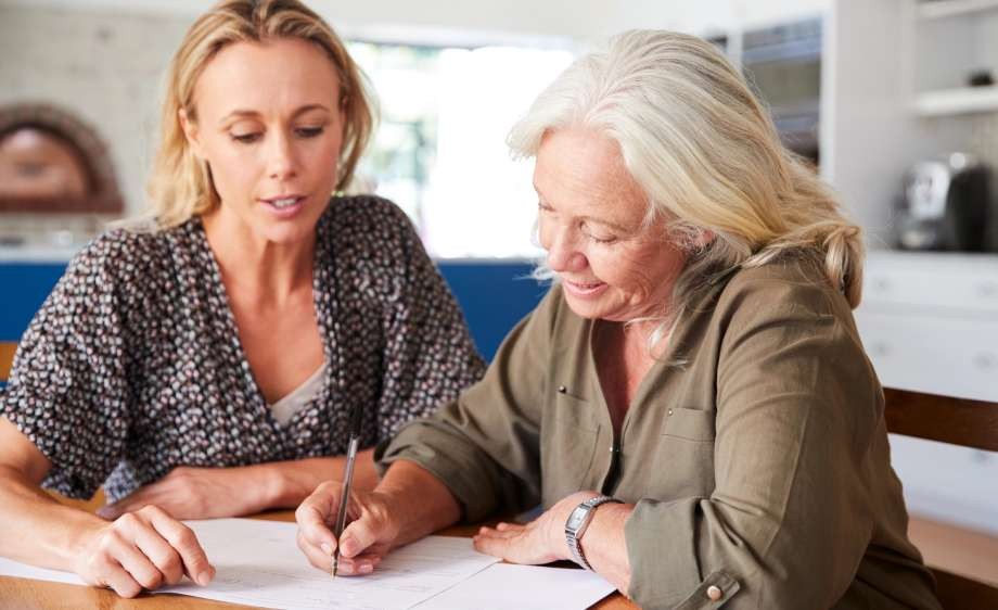 An older woman signing documents while a younger woman assists her.
