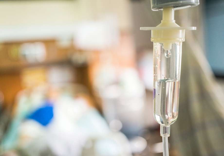 Close-up of an IV drip chamber with a blurred patient in a hospital room.