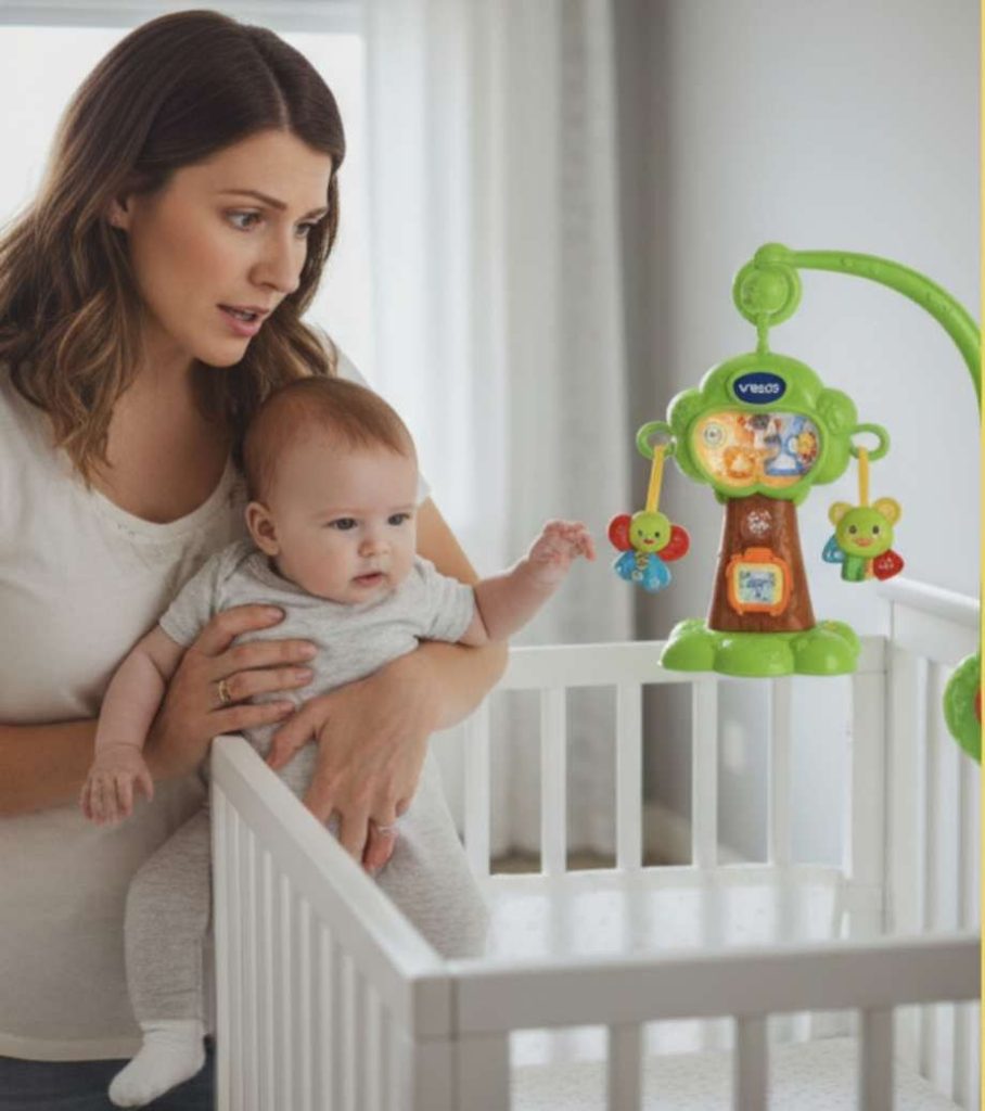 Mother holding infant baby while looking at colorful crib mobile toy.