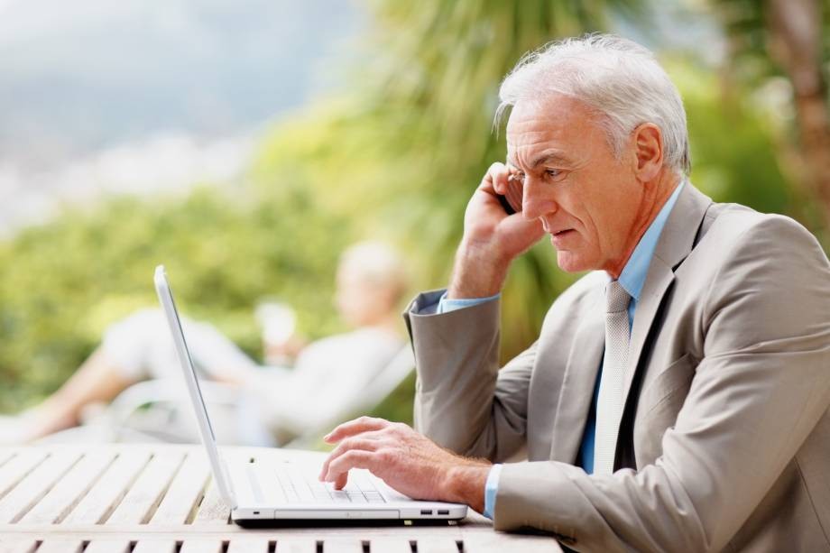 A senior man in a grey suit works on a laptop while talking on a mobile phone outdoors.