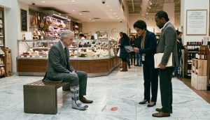 An older man, an injured patron who slipped on prosciutto, sits with a medical boot on a deli bench in Brooklyn, NYC as two professionals stand before him, one holding documents in the brightly lit store.
