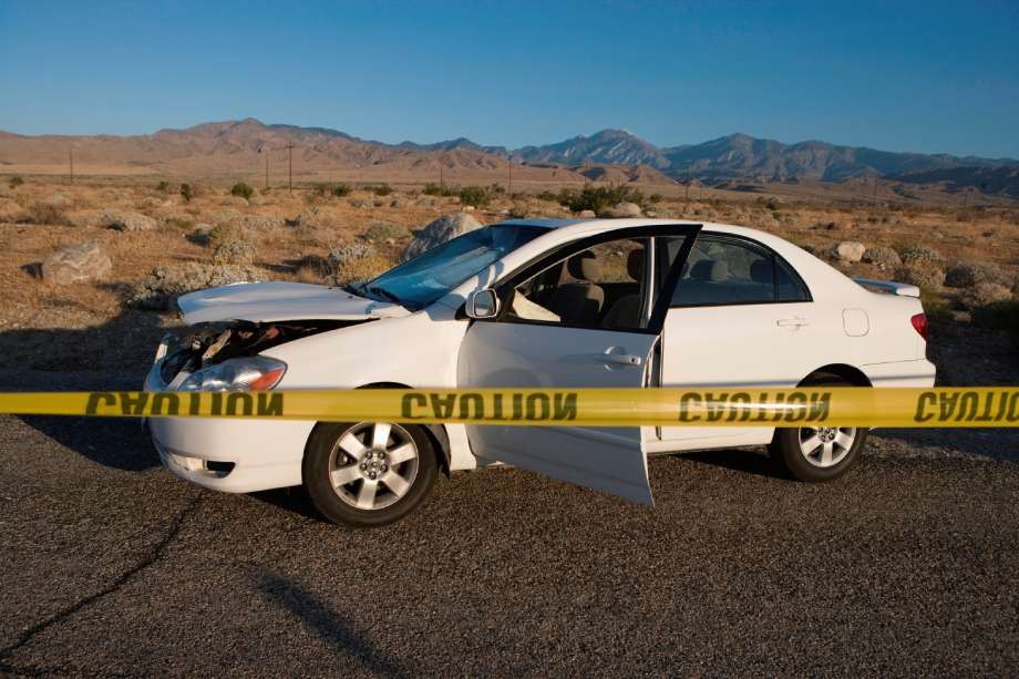A white car with a damaged front end is parked on a desert road, its driver door open and yellow caution tape in front—reminiscent of a Bensonhurst accident following a medical episode in Brooklyn, NYC.