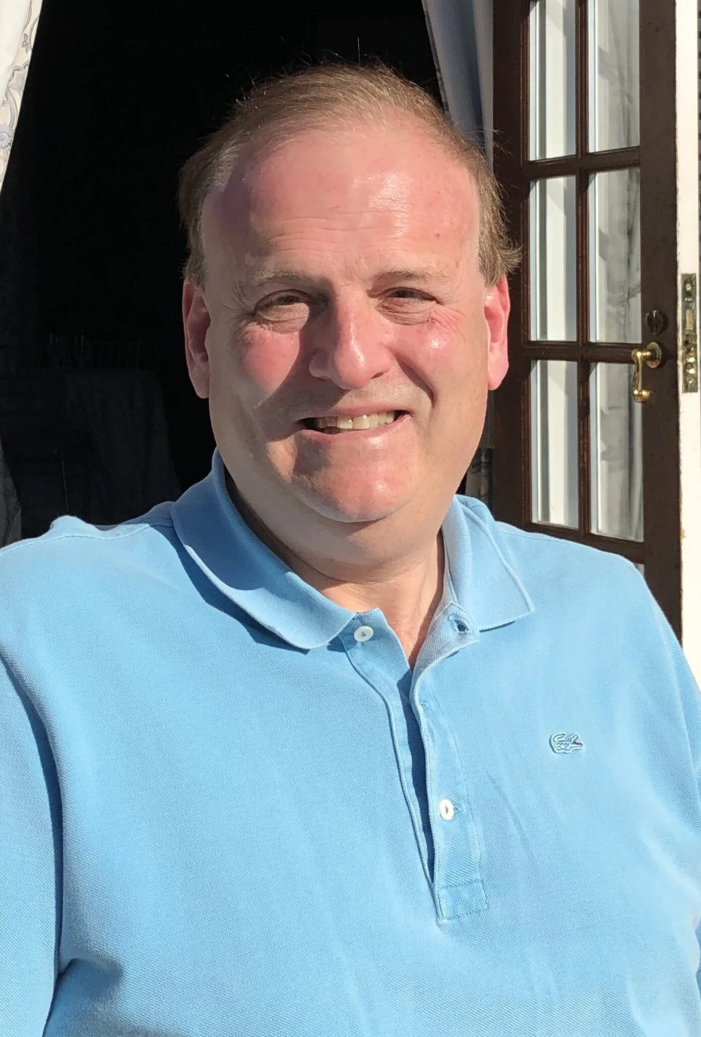 A middle-aged man wearing a light blue polo shirt sits outdoors in sunlight, smiling, with a door and white curtain in the background.