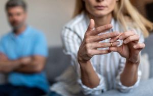 A woman removes a wedding ring from her finger while a man sits in the background, arms crossed, both appearing serious—raising the question: How Does Cheating Affect a Divorce Case?.
