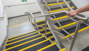 A person holds a stainless steel handrail on a concrete stairwell with yellow safety strips and non-slip black surfaces, highlighting measures that address being liable for stairway slips and falls. Emergency exit signs are visible on the walls.
