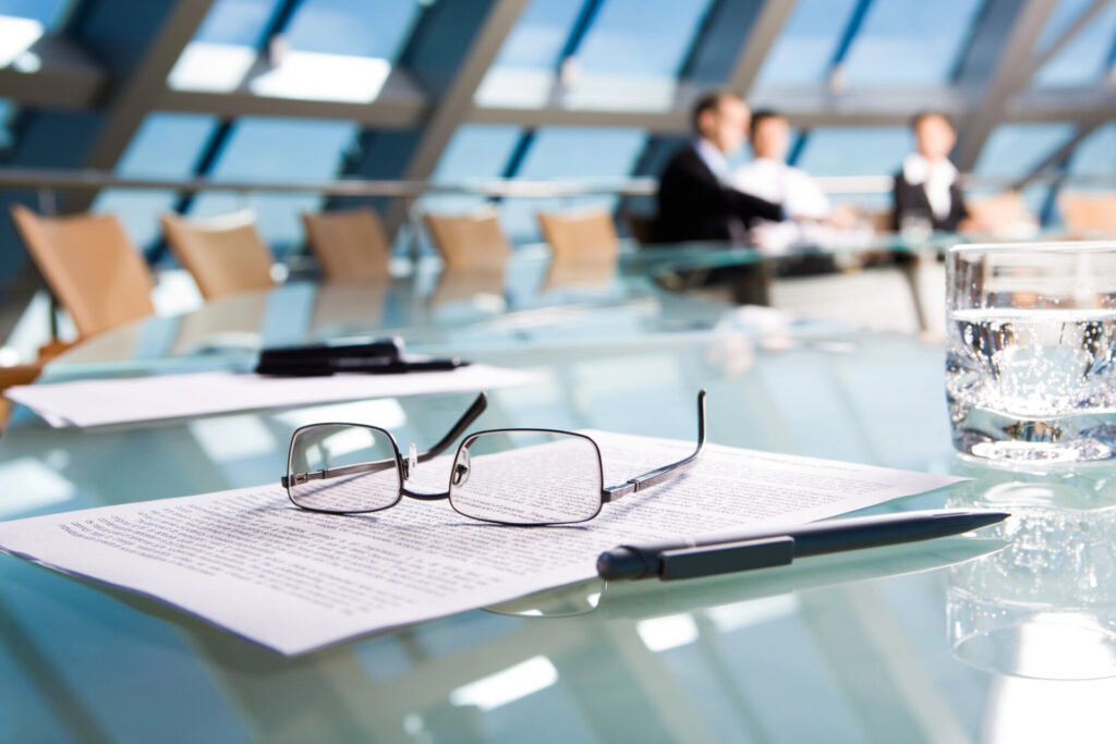 Glasses, a pen, and documents rest on a glass conference table with a glass of water; three people are blurred in discussion in the background, reflecting the professional clarity of Homepage V2-old.