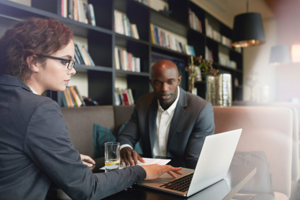 Two people in business attire sit at a table in a modern office space with bookshelves, discussing work with a laptop and documents for the Homepage V2-old project.