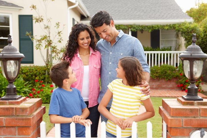 Happy family of four standing together behind white suburban picket fence.