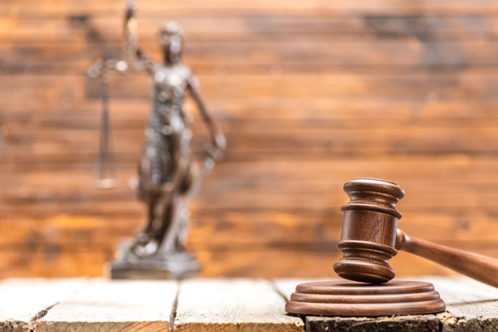Wooden judge gavel on desk with Lady Justice statue in background.