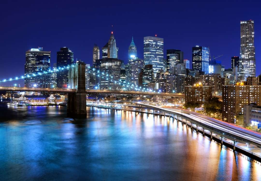 Night view of city skyline locations with illuminated skyscrapers, a lit suspension bridge, and traffic along a waterfront highway reflected in the river.