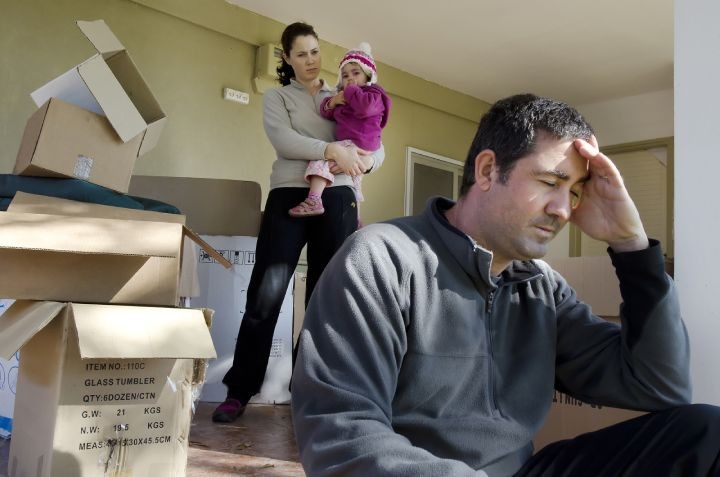 Stressed man sitting near moving boxes with woman and child behind.