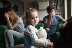 Sad young girl holding teddy bear with stressed parents in background.