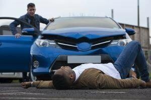 Injured pedestrian lying on the road in front of a damaged blue car.
