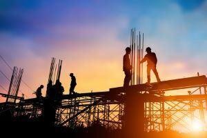 Construction workers stand and work on scaffolding at a building site during sunset, raising the question: Who Is Liable for Injuries in a New York Construction Accident? Metal rods and a colorful sky frame the scene.