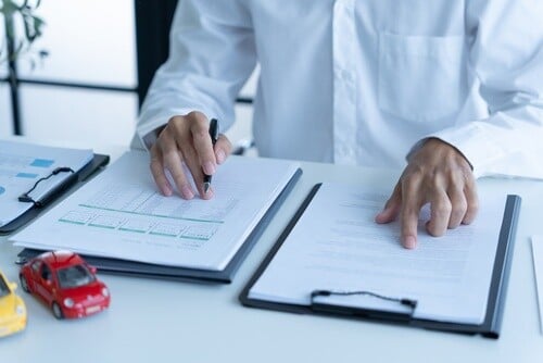 Person signing insurance forms beside toy cars and clipboards on desk