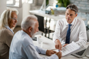 A man in a suit explains paperwork to an older couple sitting across the table from him in a bright office setting.