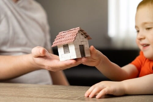 An adult and a child hold a small model house together on a wooden table.