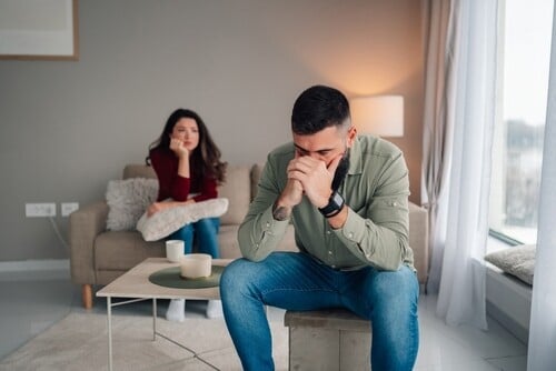 A man sits on a stool looking distressed, with his hands clasped near his face, while a woman sits on a couch in the background, watching him thoughtfully.
