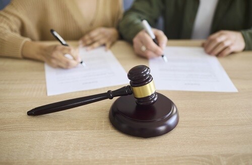 two individuals signing legal documents at desk with judge gavel foreground