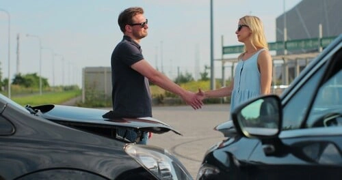 A man and woman shake hands in front of two cars that appear to have been in a minor collision, with both vehicles' hoods open and damage visible.